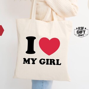 May include: Cream-colored tote bag with the text "I ❤️ MY GIRL" in black and red. The bag is held by a person wearing a cream-colored sweater. The word "NATURAL" is printed on a cream-colored circle.