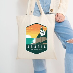 Op de afbeelding: Een witte tote bag met een groen en oranje ontwerp met een vuurtoren en de tekst "Acadia National Park".