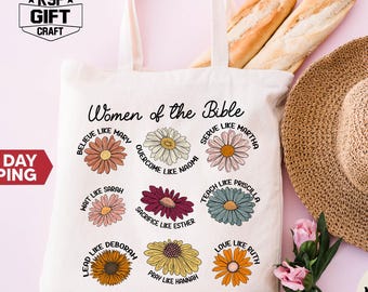 May include: Cream-colored tote bag with the text "Women of the Bible" and floral designs. The bag features various phrases like "Believe like Mary" and "Love like Ruth." A straw hat and two baguettes are in the background.