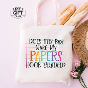 May include: A white canvas tote bag with the text "Does this bag make my papers look graded?" in rainbow colors. The text is on a blue and white lined background, mimicking notebook paper.