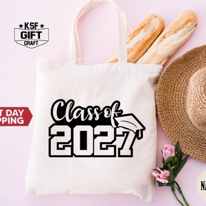 May include: A natural-colored canvas tote bag featuring the black printed text Class of 2027 with a graduation cap icon. The bag is displayed alongside two baguette-style loaves of bread, a woven straw hat, and a small pink flower stem.