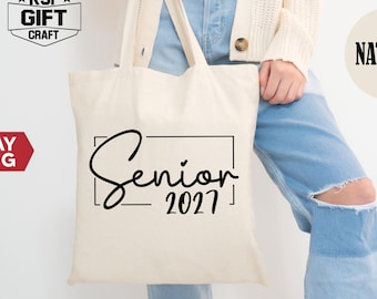 Puede incluir: Una bolsa de tela de color natural con el texto negro Senior 2027 encerrado en un marco rectangular. La bolsa tiene dos asas para el hombro y está hecha de una tela de color claro. El diseño está centrado en la parte frontal de la bolsa.