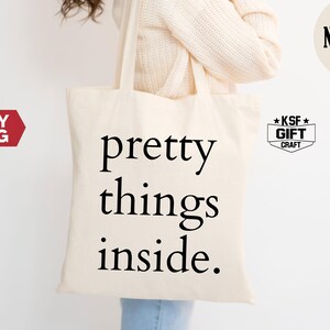 May include: Cream-colored tote bag with the words "pretty things inside" in black. The bag is held by a person wearing a cream-colored sweater. A circular "NATURAL" logo and a "KSF GIFT CRAFT" logo are visible.