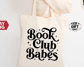 May include: A natural canvas tote bag with black text that reads "Book Club Babes" in a retro font. The bag is being held by a person.