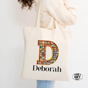 May include: A white canvas tote bag with a large letter "D" made of bookshelves filled with colorful books. The name "Deborah" is printed below the letter. The bag has a black strap.