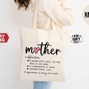 May include: Cream-colored tote bag with the word "mother" in black script and a pink heart. Below, a definition of "mother" is printed in black text. The bag is held by a person wearing a cream-colored sweater. The word "NATURAL" is printed on a cream-colored circle.