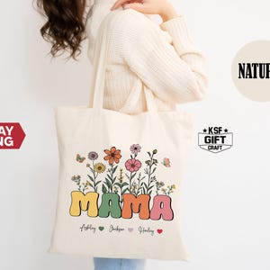 May include: Cream-colored tote bag with a floral design and the word "MAMA" in colorful bubble letters. The bag features a variety of flowers and butterflies, with names and hearts below. The bag is made of a natural material.