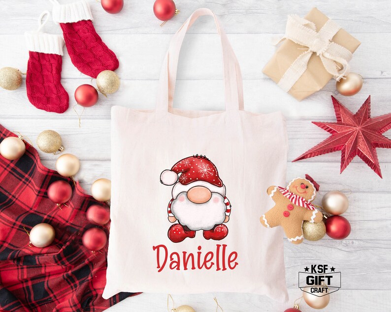 May include: A white canvas tote bag with a red and white gnome illustration and the name "Danielle" printed in red. The bag is surrounded by Christmas decorations, including red and white stockings, red ornaments, and a red star.