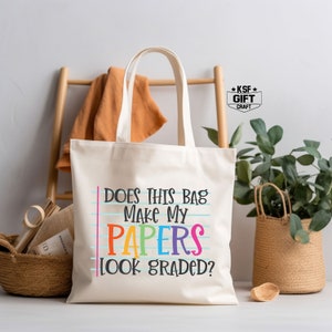 May include: A white canvas tote bag with the text "Does this bag make my papers look graded?" in rainbow colors. The text is on a blue and white lined background, mimicking a notebook page.