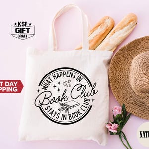 May include: A cream-colored tote bag with the text "WHAT HAPPENS IN Book Club STAYS IN BOOK CLUB" in a black circular design. The bag is next to a straw hat, two baguettes, and pink flowers. The bag has a natural color.