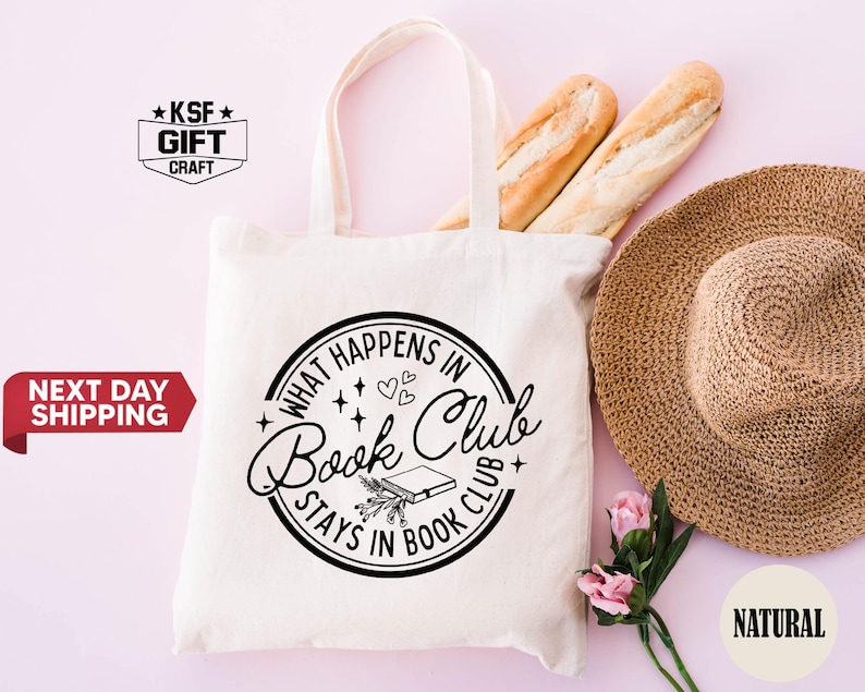 May include: A cream-colored tote bag with the text "WHAT HAPPENS IN Book Club STAYS IN BOOK CLUB" in a black circular design. The bag is next to a straw hat, two baguettes, and pink flowers. The bag has a natural color.