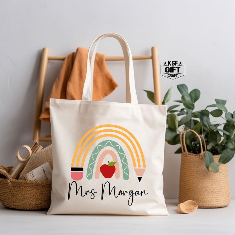 May include: A white canvas tote bag with a rainbow design and the text "Mrs. Morgan". The rainbow is made up of yellow, orange, green, and pink stripes. There are two pencils on either side of the rainbow and a red apple in the center.