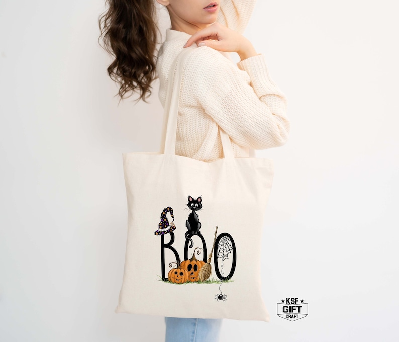 May include: A white tote bag with a black cat, pumpkins, and the word "BOO" printed on it. The cat is wearing a witch's hat and holding a broom. There is a spider web and a spider on the bag.