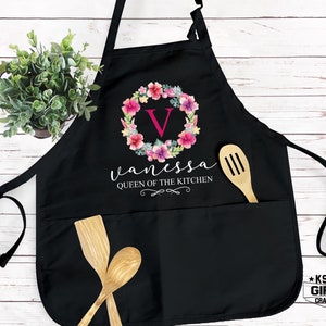 May include: Black kitchen apron with a floral wreath design and the name "Vanessa" written below it. The wreath has a pink "V" in the center. The text "Queen of the Kitchen" is written below the name.