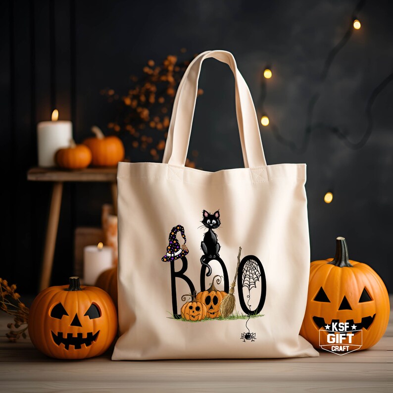 May include: A white canvas tote bag with a black cat, a witch's hat, and the word "BOO" printed on it. There are also pumpkins and a spider web on the bag. The bag is sitting on a wooden surface with pumpkins in the background.