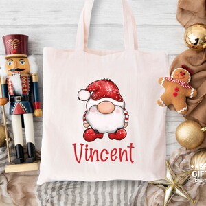 May include: A pink tote bag with a white gnome illustration wearing a red and white hat. The gnome is holding a red and white candy cane. The bag has the name "Vincent" printed in red letters.
