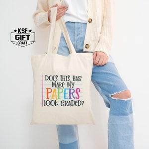 May include: A white canvas tote bag with the text "Does this bag make my papers look graded?" in black and rainbow colors. The text is on a blue and white lined background, mimicking notebook paper.