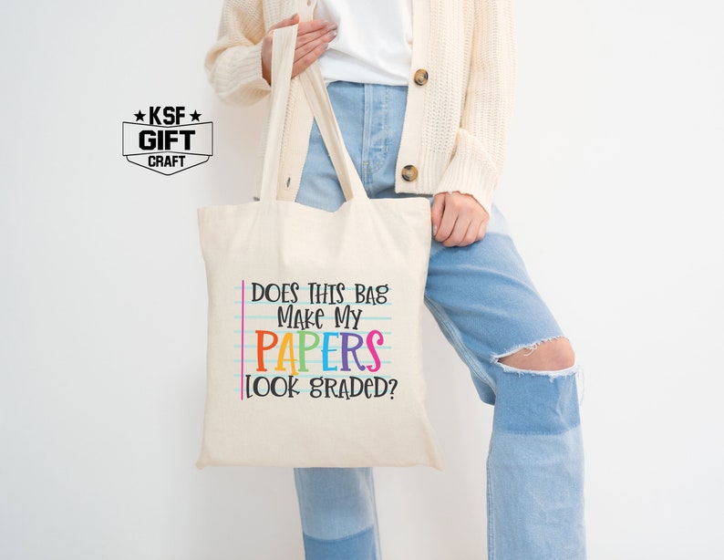 May include: A white canvas tote bag with the text "Does this bag make my papers look graded?" in black and rainbow colors. The text is on a blue and white lined background, mimicking notebook paper.