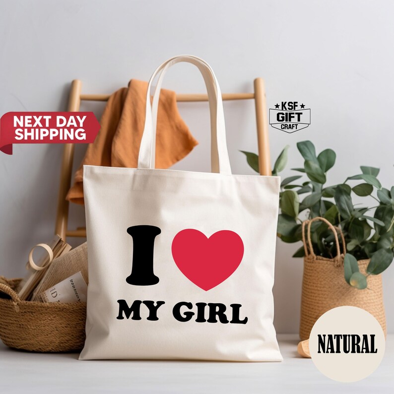 May include: Cream-colored tote bag with black text and a red heart. The text reads "I MY GIRL". The bag has long handles and is made of a sturdy fabric. A wooden ladder and greenery are in the background.