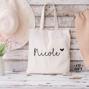 May include: White canvas tote bag with the name "Nicole" written in black cursive with a small black heart next to it. The bag has black handles and is sitting on a white wooden surface.