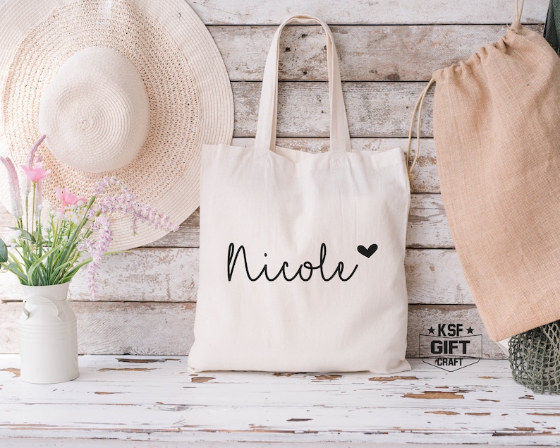 May include: White canvas tote bag with the name "Nicole" written in black cursive with a small black heart next to it. The bag has black handles and is sitting on a white wooden surface.