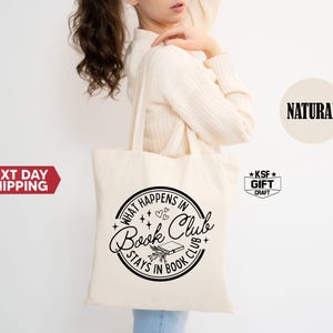 May include: A cream-colored tote bag with a black circular design that reads "What Happens in Book Club Stays in Book Club." The design includes stars, hearts, and a book illustration. The bag is held by a person.
