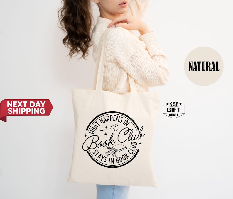 May include: A cream-colored tote bag with a black circular design that reads "What Happens in Book Club Stays in Book Club." The design includes stars, hearts, and a book illustration. The bag is held by a person.