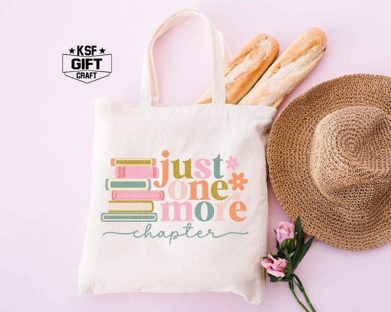 Op de afbeelding: Een witte tote bag met een kleurrijke afbeelding waarop "just one more chapter" staat en een stapel boeken. De tas heeft een bruin handvat en is gemaakt van canvas. De afbeelding is in retro-stijl met een stapel boeken en de woorden in verschillende kleuren.