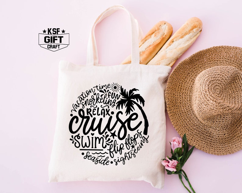May include: A white canvas tote bag with black text that says "vacation time, snorkeling, fun, relax, cruise, swim, flip flops, seaside, sightseeing". The text is arranged in a circle around a palm tree. The bag is on a pink background with a straw hat, bread, and pink flowers.