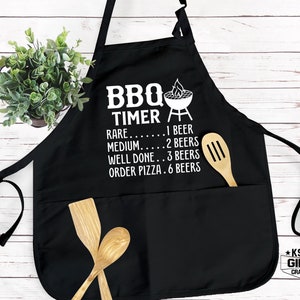 May include: Black BBQ apron with a white graphic that says "BBQ Timer" and lists the doneness of meat with corresponding beer counts. The graphic includes a grill and flames. The apron has two pockets and a wooden spoon in one of the pockets.