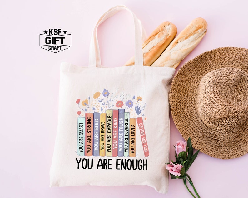 May include: A white tote bag with a design of colorful books with positive affirmations. The text on the books reads "You are smart", "You are strong", "You are bold", "You are brave", "You are capable", "You are kind", "You are tough", "You are powerful", "You are loved", "You are valued". The text at the bottom of the bag reads "You are enough".