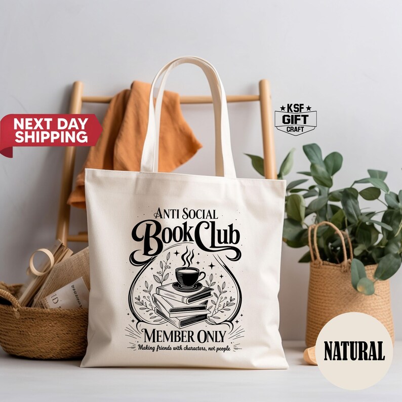 Op de afbeelding: Een natuurlijke canvas draagtas met de tekst "Anti Social Book Club" in een decoratief lettertype. Het ontwerp bevat een stapel boeken, een kopje koffie en de woorden "Member Only". De tas heeft een eenvoudige handgreep en wordt getoond met andere decoratieve items.