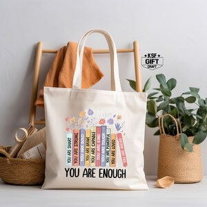 May include: A white canvas tote bag with a design of colorful books stacked on top of each other. The books have words printed on them, such as "You are smart", "You are strong", "You are bold", "You are brave", "You are capable", "You are kind", "You are tough", "You are powerful", "You are loved", "You are valued". The words are printed in a rainbow of colors. The design is surrounded by flowers. The tote bag has the words "You are enough" printed below the design.