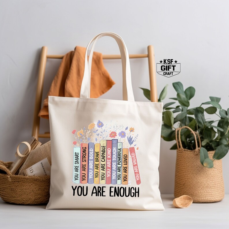 May include: A white canvas tote bag with a design of colorful books stacked on top of each other. The books have words printed on them, such as "You are smart", "You are strong", "You are bold", "You are brave", "You are capable", "You are kind", "You are tough", "You are powerful", "You are loved", "You are valued". The words are printed in a rainbow of colors. The design is surrounded by flowers. The tote bag has the words "You are enough" printed below the design.