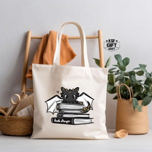 Pu&ograve; includere: Una borsa tote in tela bianca con un'illustrazione di un drago nero e dorato. Il drago &egrave; seduto su una pila di libri con il testo "book dragon" sotto. La borsa ha manici neri e il testo "KSF GIFT CRAFT" in alto a destra.