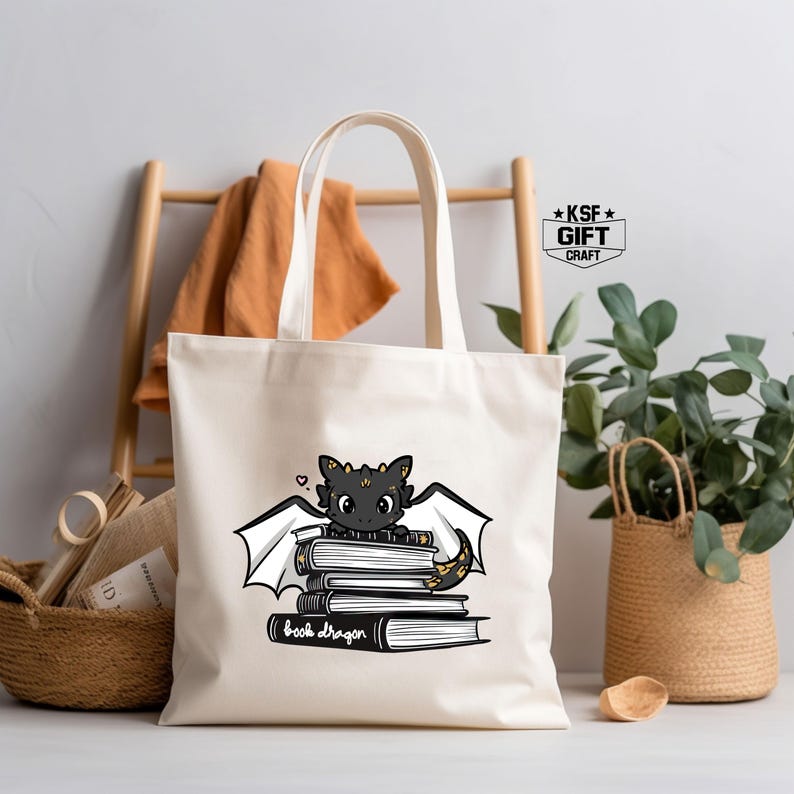 Pu&ograve; includere: Una borsa tote in tela bianca con un'illustrazione di un drago nero e dorato. Il drago &egrave; seduto su una pila di libri con il testo "book dragon" sotto. La borsa ha manici neri e il testo "KSF GIFT CRAFT" in alto a destra.
