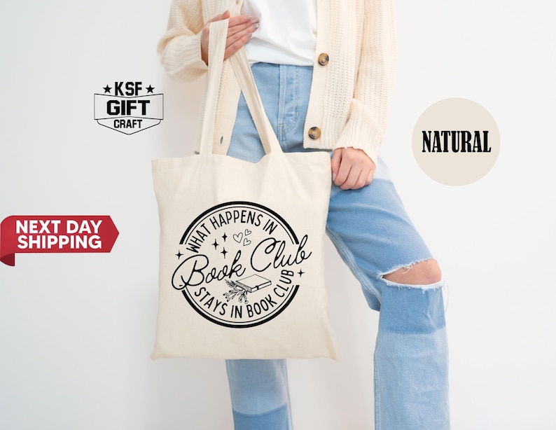 May include: A cream-colored tote bag with black text that reads "What happens in Book Club stays in Book Club." The bag has a circular design with a book and floral elements. The bag is held by a person wearing a cream-colored cardigan and blue jeans.