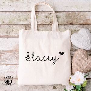 May include: A white canvas tote bag with the name "Stacey" written in black cursive script and a small black heart. The bag is sitting on a wooden surface with two decorative hearts.
