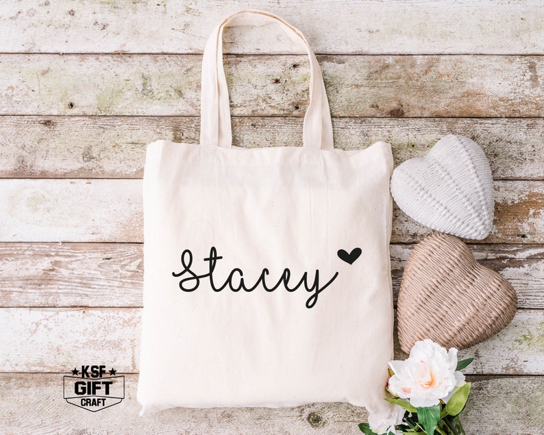 May include: A white canvas tote bag with the name "Stacey" written in black cursive script and a small black heart. The bag is sitting on a wooden surface with two decorative hearts.