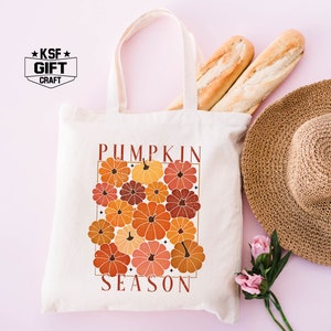 May include: A white tote bag with a design of orange, red, and brown pumpkins with the text "Pumpkin Season" in brown lettering. The bag has a brown handle and is on a pink background.