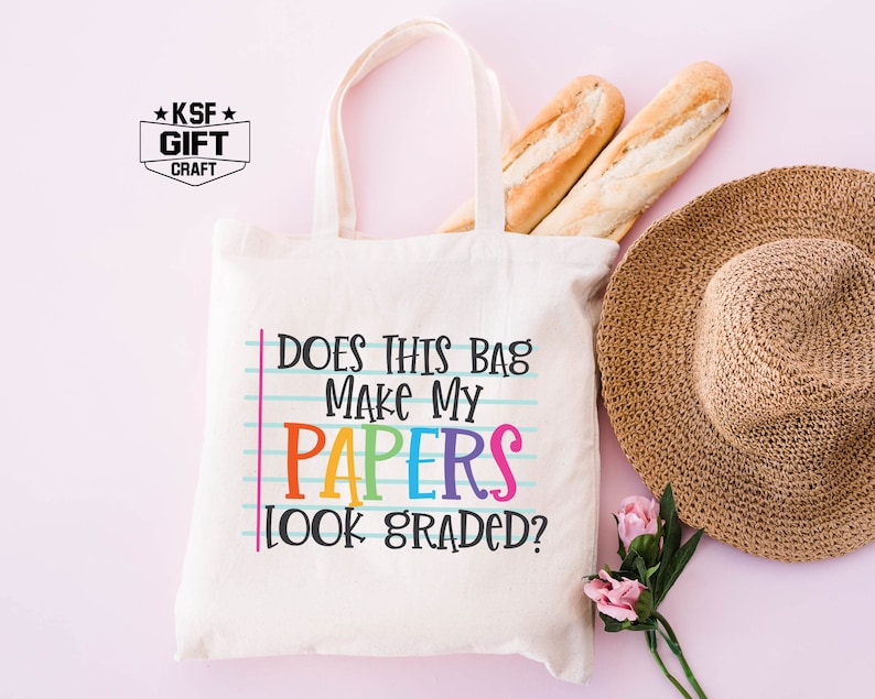 May include: A white canvas tote bag with the text "Does this bag make my papers look graded?" in rainbow colors. The text is on a blue and white lined background, mimicking notebook paper.