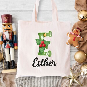 May include: A pink canvas tote bag featuring a green letter E adorned with holly and snowflakes. The name Esther is written in black cursive beneath the letter.