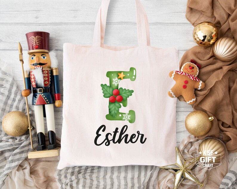 May include: A pink canvas tote bag featuring a green letter E adorned with holly and snowflakes. The name Esther is written in black cursive beneath the letter.