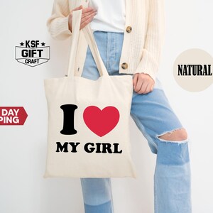 May include: Cream-colored tote bag with the text "I ❤️ MY GIRL" in black and red. The bag is held by a person wearing a cream-colored cardigan and blue jeans.