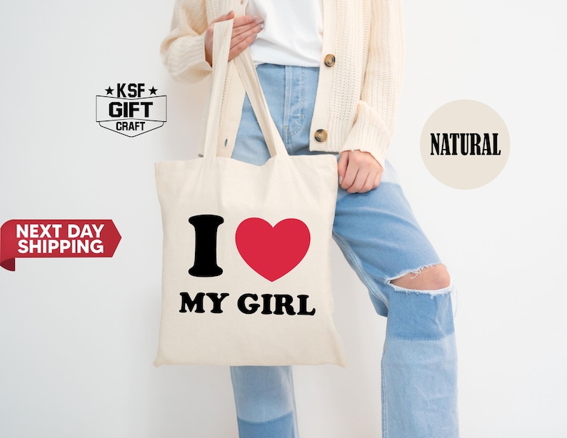 May include: Cream-colored tote bag with the text "I ❤️ MY GIRL" in black and red. The bag is held by a person wearing a cream-colored cardigan and blue jeans.