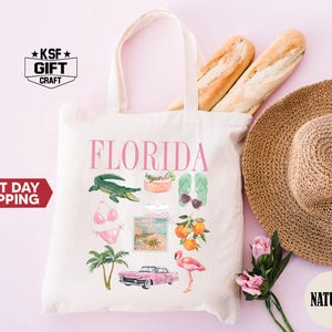 May include: A white tote bag with a pink and green design featuring the word "FLORIDA" and images of a flamingo, palm trees, a cocktail, sunglasses, a bikini, and an alligator. The bag has natural fabric handles.
