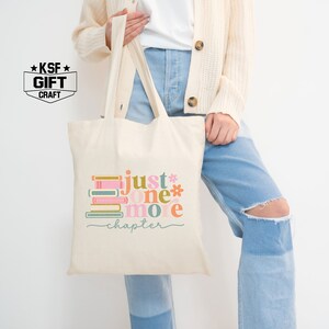 Op de afbeelding: Een witte canvas tote bag met een kleurrijke afbeelding van gestapelde boeken en de tekst "just one more chapter".