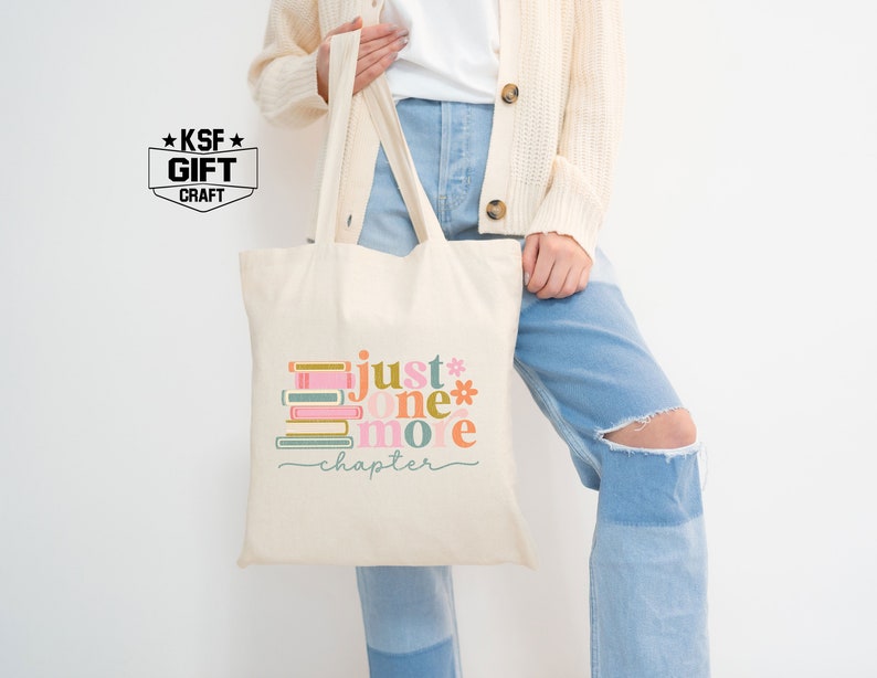 Op de afbeelding: Een witte canvas tote bag met een kleurrijke afbeelding van gestapelde boeken en de tekst "just one more chapter".