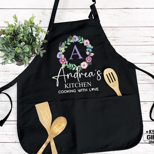 May include: Black kitchen apron with a floral wreath design and the text "Andrea's Kitchen Cooking with Love". The wreath has a purple "A" in the center.