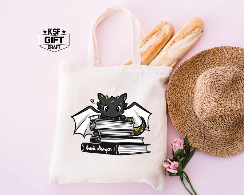Pu&ograve; includere: Una borsa tote bianca con manici neri presenta un drago di cartone animato nero e oro seduto su una pila di libri. Il testo "book dragon" &egrave; stampato sotto il drago. La borsa ha un piccolo logo bianco e nero che dice "KSF GIFT CRAFT" nell'angolo in alto a sinistra.